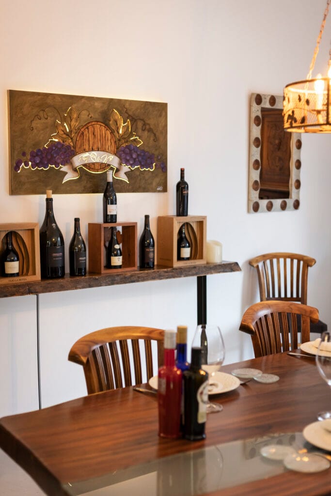 Detalle del bar de vinos con botellas expuestas sobre una tabla de madera y arte temático de uvas y bodega.