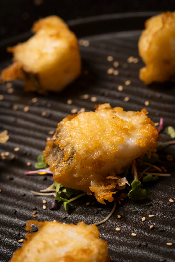 Primer plano de un trozo de pescado fresco rebozado y crujiente, servido sobre brotes verdes y semillas de sésamo.